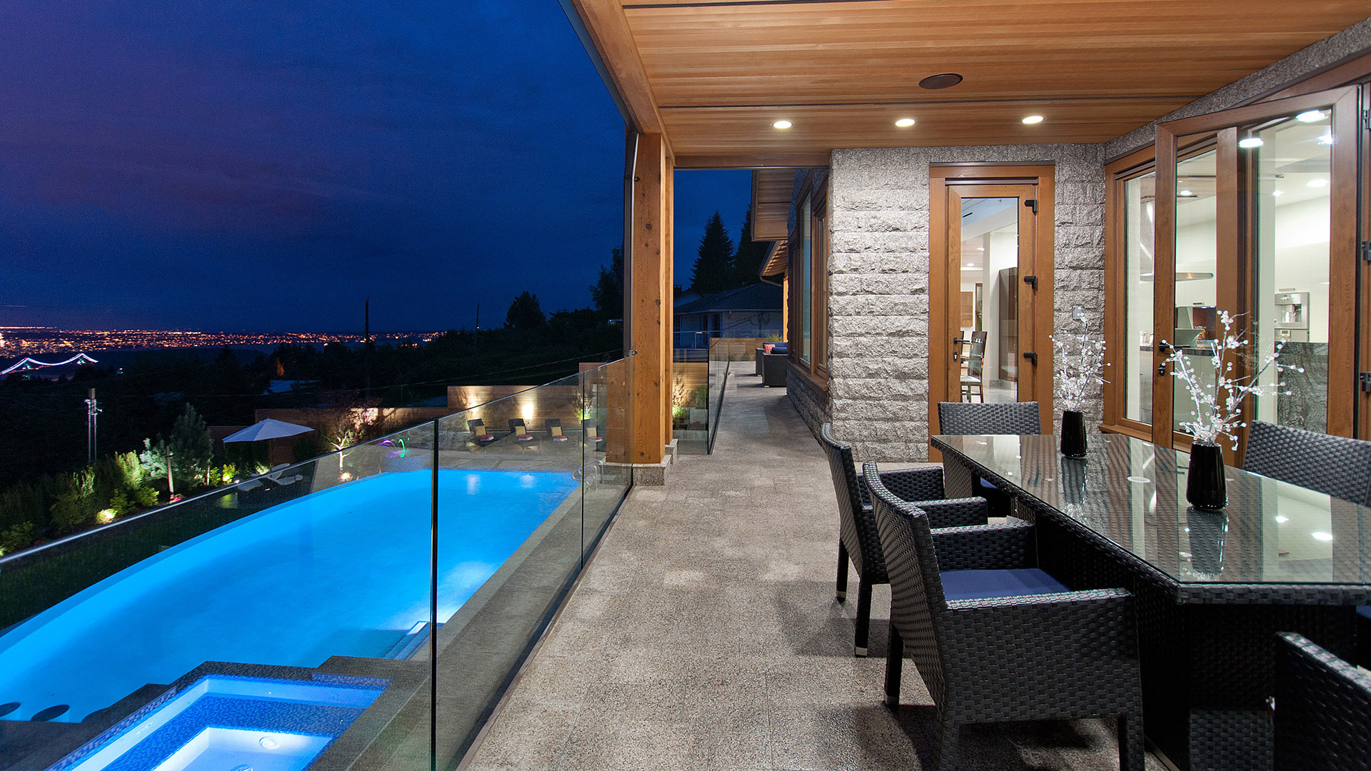 Covered deck at twilight with pool view glass railings granite columns and outdoor dining with city lights Millstream British Properties West Vancouver