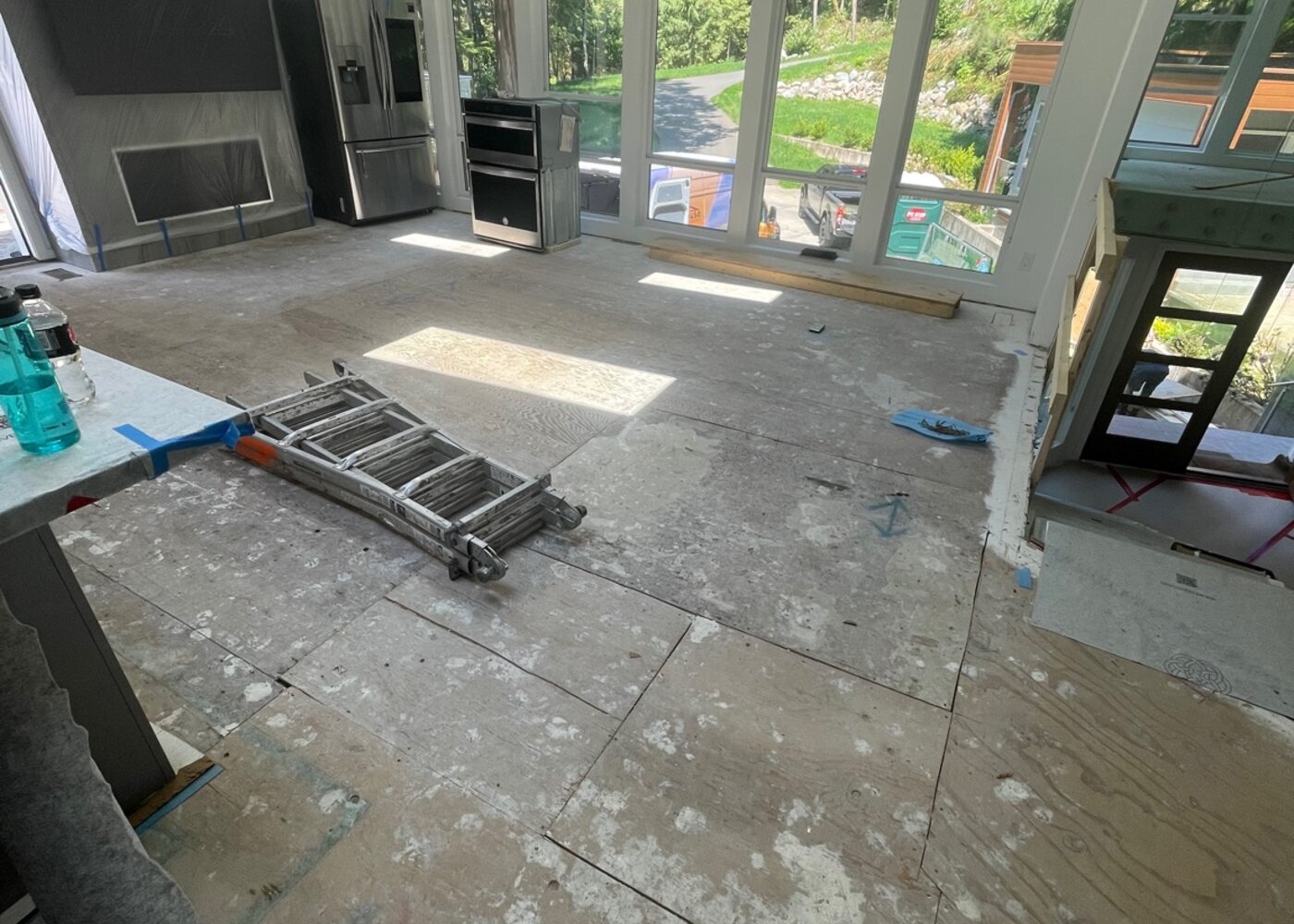 Open-concept kitchen and living area stripped to subfloor during insurance restoration with ladder, appliances, and floor-to-ceiling windows visible — North Vancouver luxury home