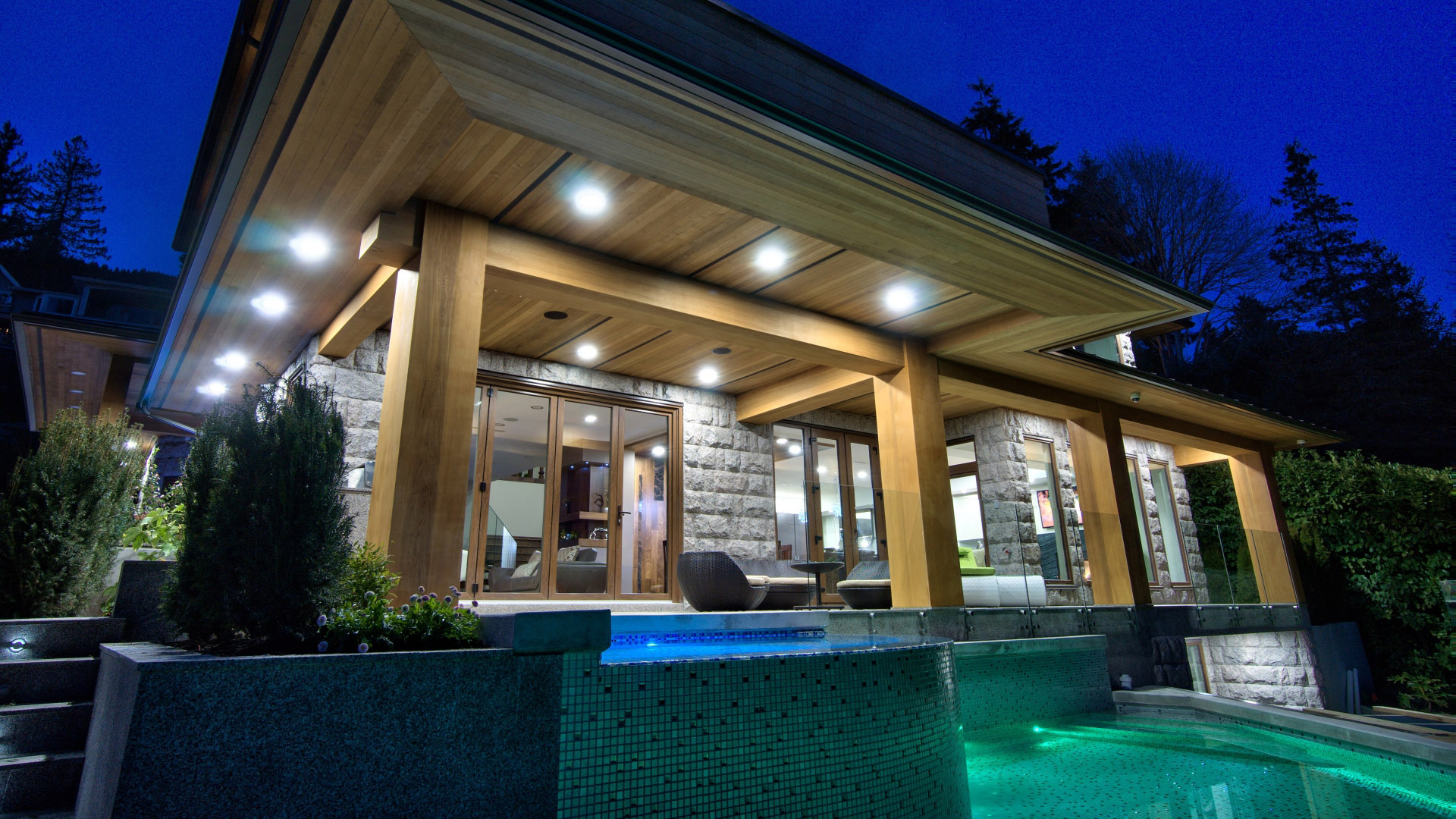Covered deck at dusk with heavy timber columns granite pillars and glass-tiled pool at West Bay luxury home West Vancouver