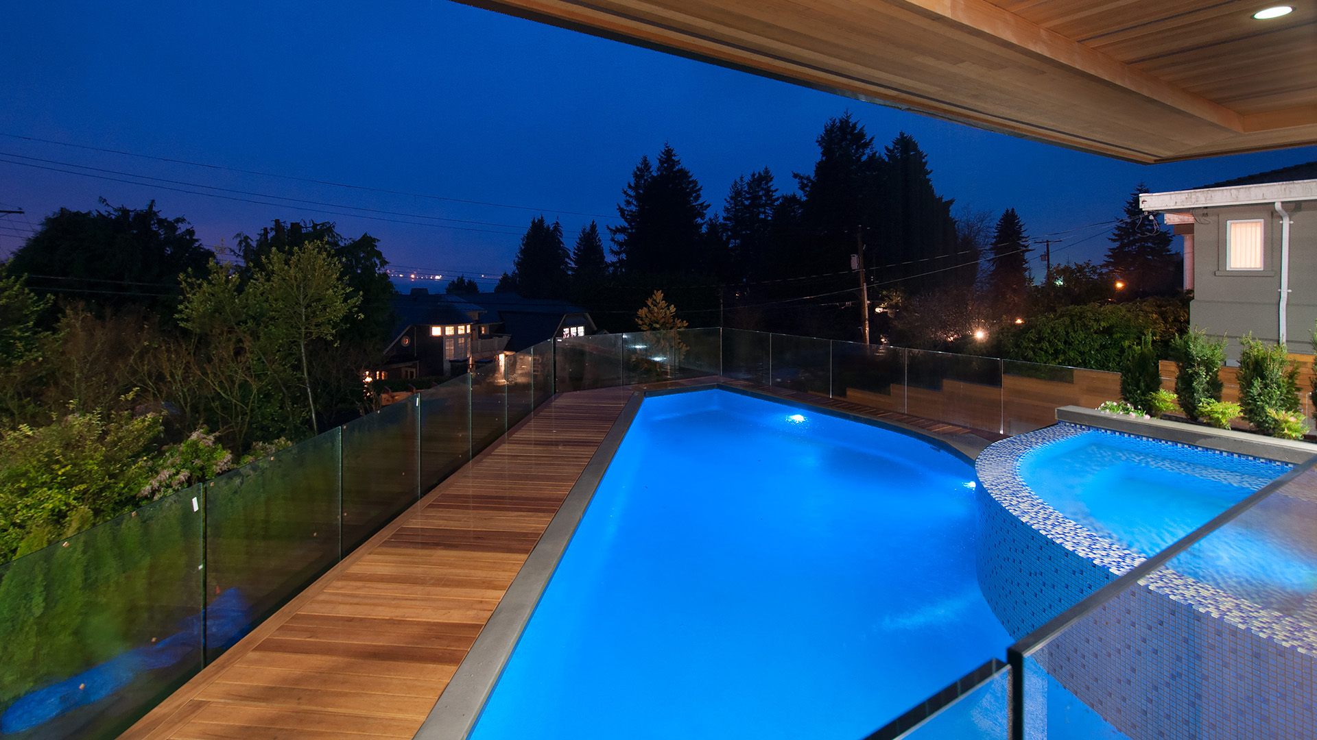 Glass-tiled swimming pool and hot tub at dusk with seamless glass railings and teak decking West Bay West Vancouver